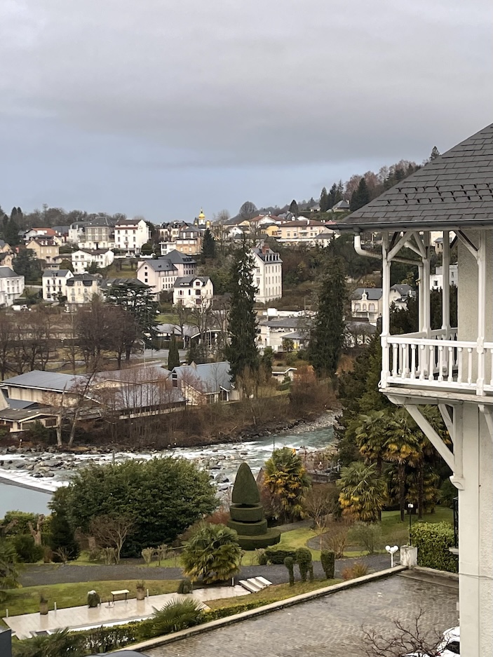 A landscape picture of the mountainous village of Lourdes, France with a fast current river running through and buildings built up along the hillside