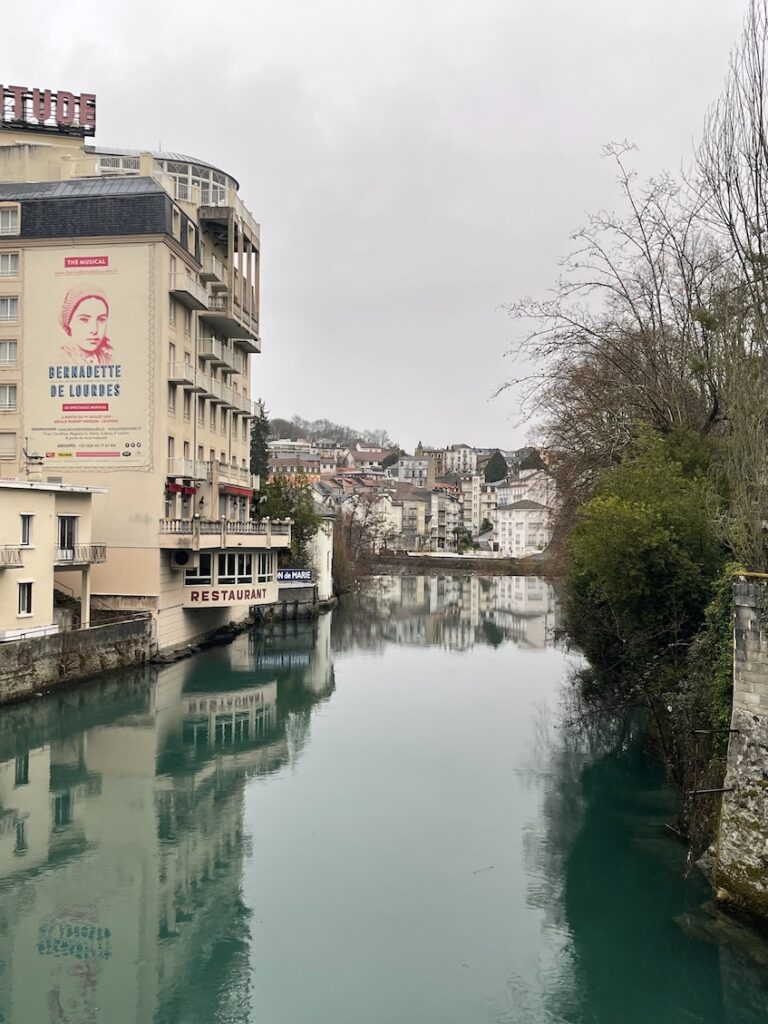 The Gave du pau river with its stunning blue water running through a village with buildings such as hotels built right up to the edge of the water and a bridge and more buildings in the background