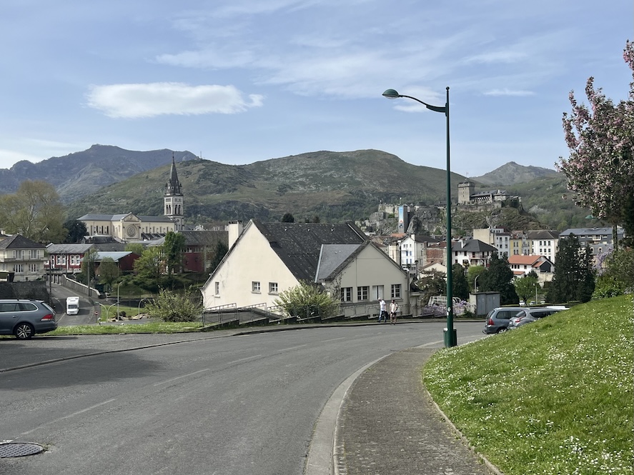 A road leading to the moutainous village of Lourdes, France with buildings and houses along the bottom and grassy hills in the backdrop