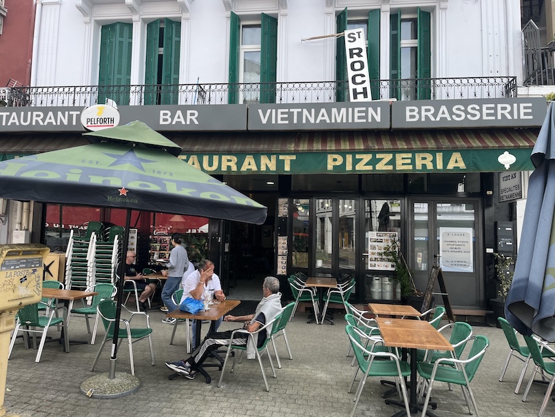 Brasserie restaurant in Lourdes, France with green shade awnings and above it is a white building with green shades, there are tables and chairs set out in front with patrons sitting at a few of the tables