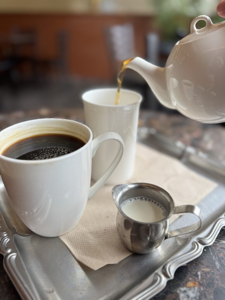 A teapot is pouring tea into a white mug and on the same silver tray is a mug of coffee and a small silver pitcher of cream