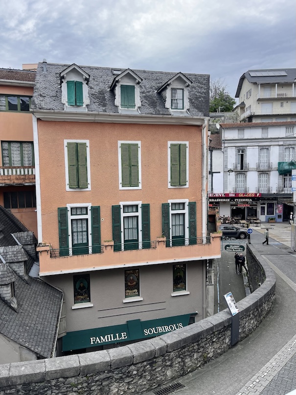 A French mountainous house in Lourdes, France with green shutters against a peach building belonging to the family Soubirous