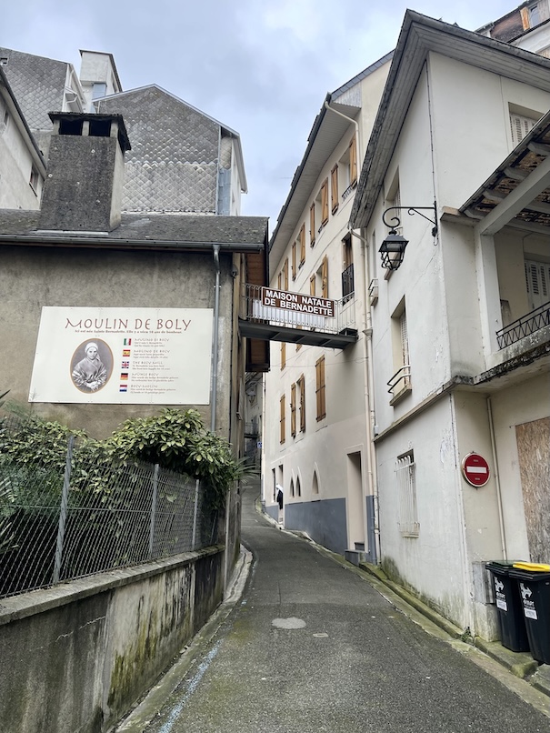 A road running up between two houses in the mountainous region of Lourdes with a sign on the houses saying birthplace of St. Bernadette