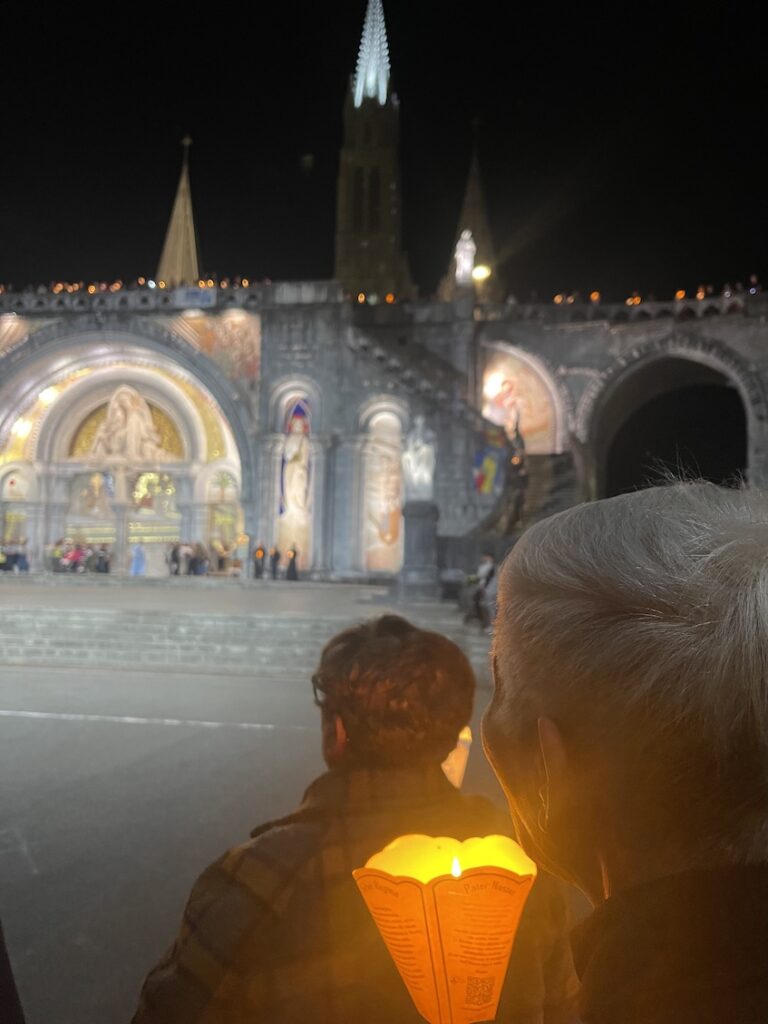 Nightly candlelight procession at lourdes with men holding candles lit inside paper cones looking at the front of a lighted up basilica at night