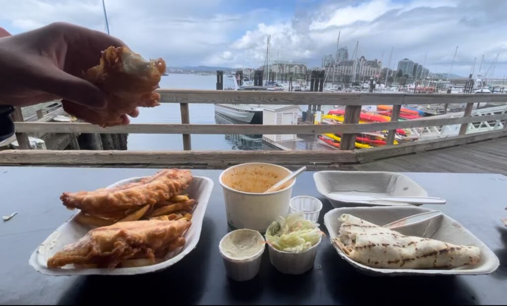 A plate of fish and chips and a plate of a wrap are on a table that looks out towards the Victoria inner harbor. A hand is holding one of the fried fish with a bite taken from it. In the middle of both plates is a cup of chowder and a small container of coleslaw and tarter sauce