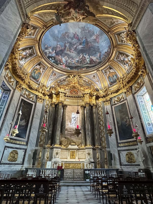 Baroque interior of the Lady Chapel featuring an ornate altar, statue of Mary flanked by columns, and a domed ceiling with a fresco of the Assumption of Mary by François Lemoyne, lit by natural light from concealed windows. Seven red tabernacle lights hang from the ceiling