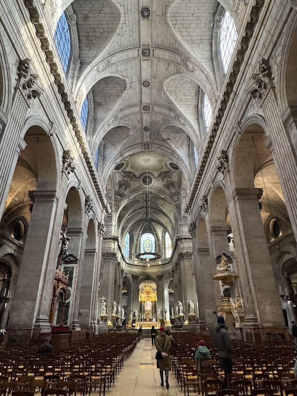 The nave of St Sulpice church, large stone columns line the sides, great windows from above drown the church with light, the center aisle leads to the main altar in the background