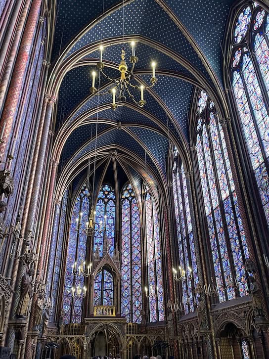 A side angle of the grandiose interior of Sainte Chapelle. lights hang from the ornate ceilings, grand stained glass windows all around