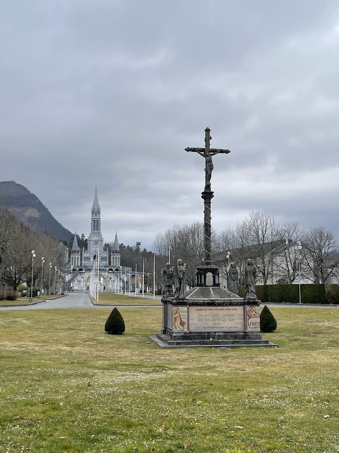 Sanctuary of Our Lady of Lourdes taken from the very front of the sanctuary which starts with a large crucifix of Our Lord and a long stretch of grass and roads leading to the basilicas far in the back on a cloudy and gloomy day