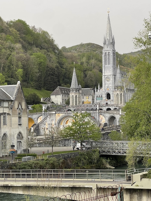 Sanctuary of Our Lady of Lourdes, France taken from a distance with the basicilas nestled between green trees and a river running under two bridges to the right of the basilicas