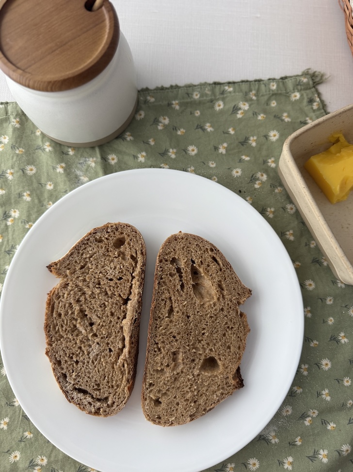 Two slices of whole wheat sourdough bread on a white plate with a green cotton napkin underneath with daisies on it. On the side is a white ceramic honey pot and to the right is a ceramic butter dish with a knob of butter inside