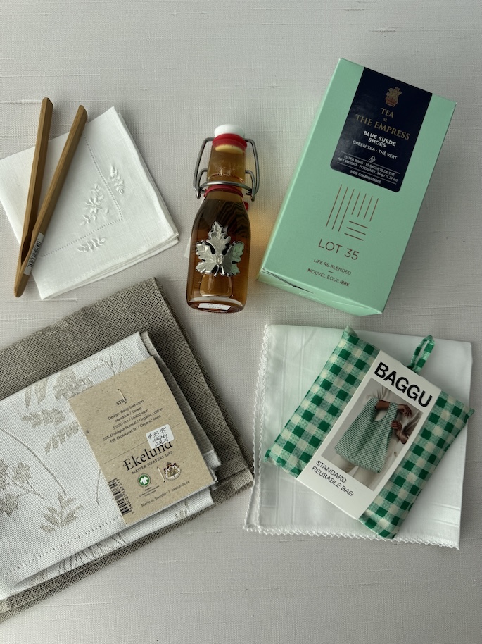 Souvenirs from Victoria, BC laid out on top of a white table with items such as a box of tea, wooden toast tongs, linen tea towels, a small maple swig top container, a Baggu reusable bag on top of cotton handkerchiefs
