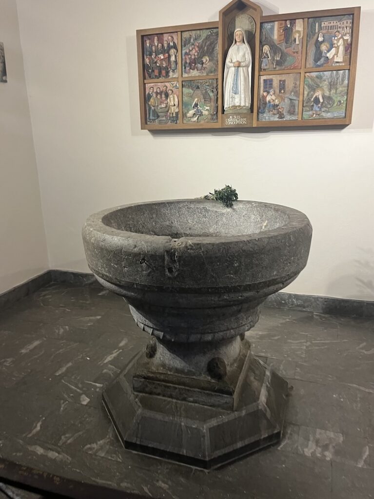 Baptismal font made of stone before icons of St. Bernadette and images of her life and the apparitions of Our Lady of Lourdes