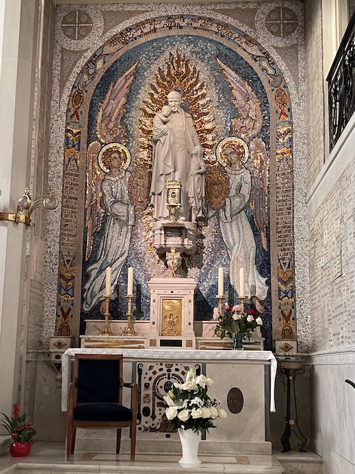 A blue wooden chair in front of a side altar, a reliquary above the altar with a mosaic of St Vincent de Paul and two angels