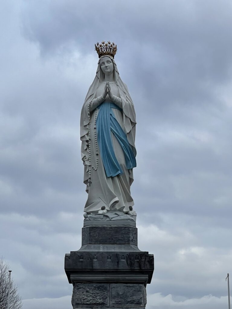 Statue of Our Lady of Lourdes that stands towering on a pillar against a cloudy gray sky