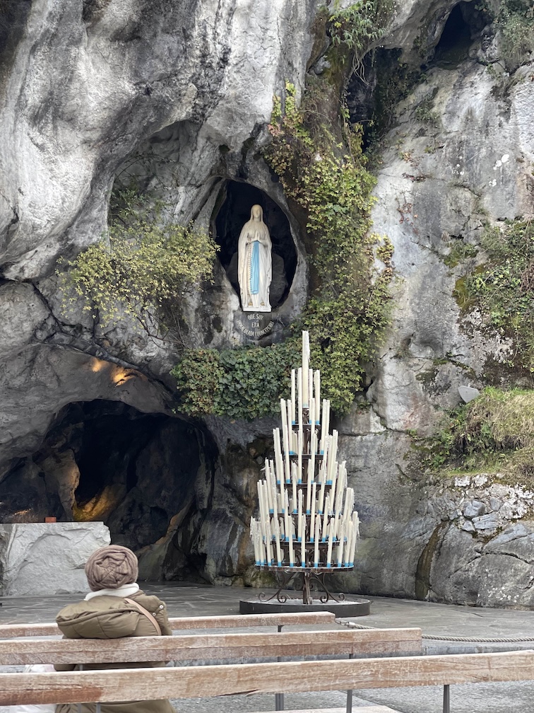 A statue of Mary inside the grotto of Massabielle in Lourdes, France, with dozens of candles. Benches int he forefront for pilgrims to pray.