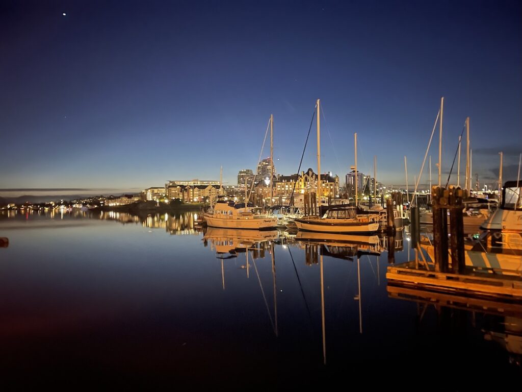 Victoria’s Inner Harbour at sunset, with glass-like waters reflecting the deep, dark blue sky. The calm surface mirrors the silhouettes of historic buildings, docked boats, and soft evening lights along the waterfront. The scene is tranquil and moody, with rich twilight tones creating a serene, reflective atmosphere