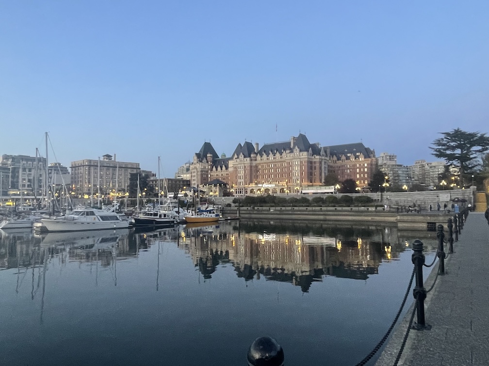 A wide view of the Fairmont Empress Hotel from across Victoria’s Inner Harbour. The iconic brick building stands majestically with its grand, château-style architecture, framed by trees and marina boats in the foreground. The still harbor water reflects the hotel and surrounding lights, adding to the elegance of the scene under a dusky sky.