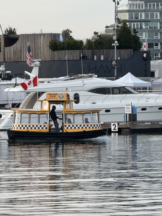 A small yellow water taxi or pickle boat glides across Victoria Harbour under a cloudy sky in the calm water. The boat’s rounded shape and bright color stand out against the backdrop of a white yacht, with heritage buildings and boats visible along the shoreline.