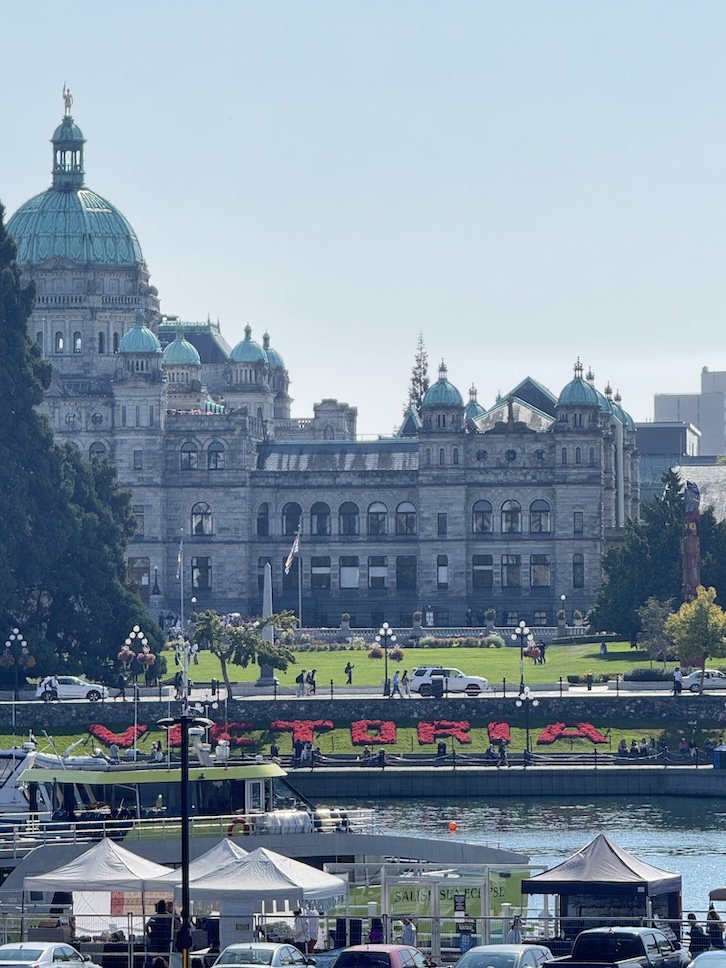 The parliament building in Victoria, BC in the background with lawns of grass before it. Right above the water on the harbor is the world "Victoria" created entirely from vibrant flowers, displayed prominently along the harbor’s edge