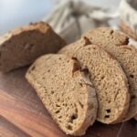 Slices of whole wheat sourdough bread on a wooden cutting board with a bread bag and basket bannetons in the background