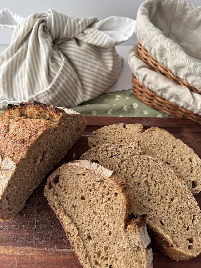 Slices and a loaf of whole wheat sourdough bread is on a cutting board with a canvas bread bag in the background and next to that are two basket bannetons stacked on top of each other