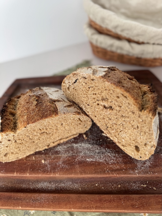 A loaf of whole wheat sourdough cut in half with one slighted leaned on top of the other on a wooden cutting board all on top of a green cotton napkin. There are 2 linen lined basket bannetons in the background