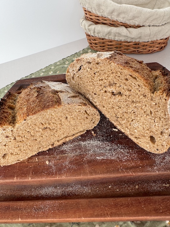 A loaf of whole wheat sourdough cut in half with one slighted leaned on top of the other on a wooden cutting board all on top of a green cotton napkin. There are 2 linen lined basket bannetons in the background