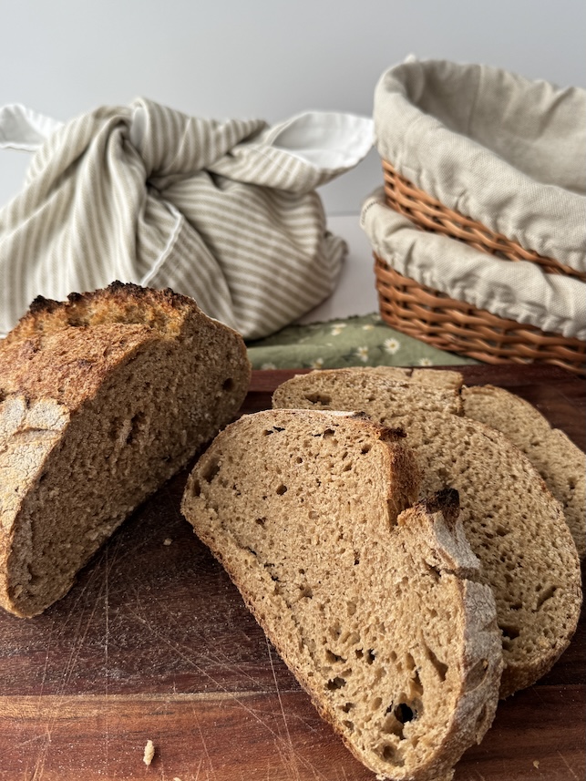 Slices and a loaf of whole wheat sourdough bread is on a cutting board with a canvas bread bag in the background and next to that are two basket bannetons stacked on top of each other