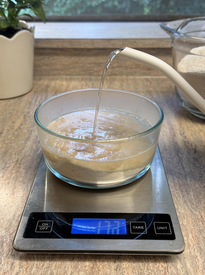 Boiling water being poured from a gooseneck kettle into a glass bowl that has flour it in. It is sitting on top of a silver kitchen scale