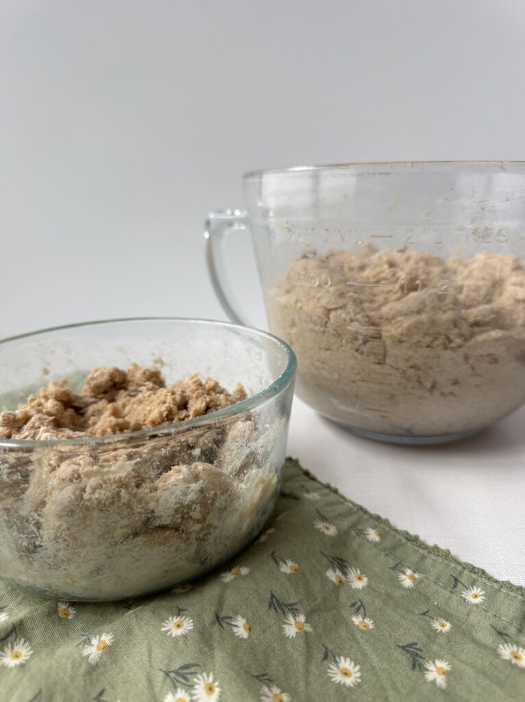 Two bowls containing dough that is being "Autolyzed". One is smaller and the other is larger with a handle. The smaller one is sitting on top of a green napkin with daisies on it.