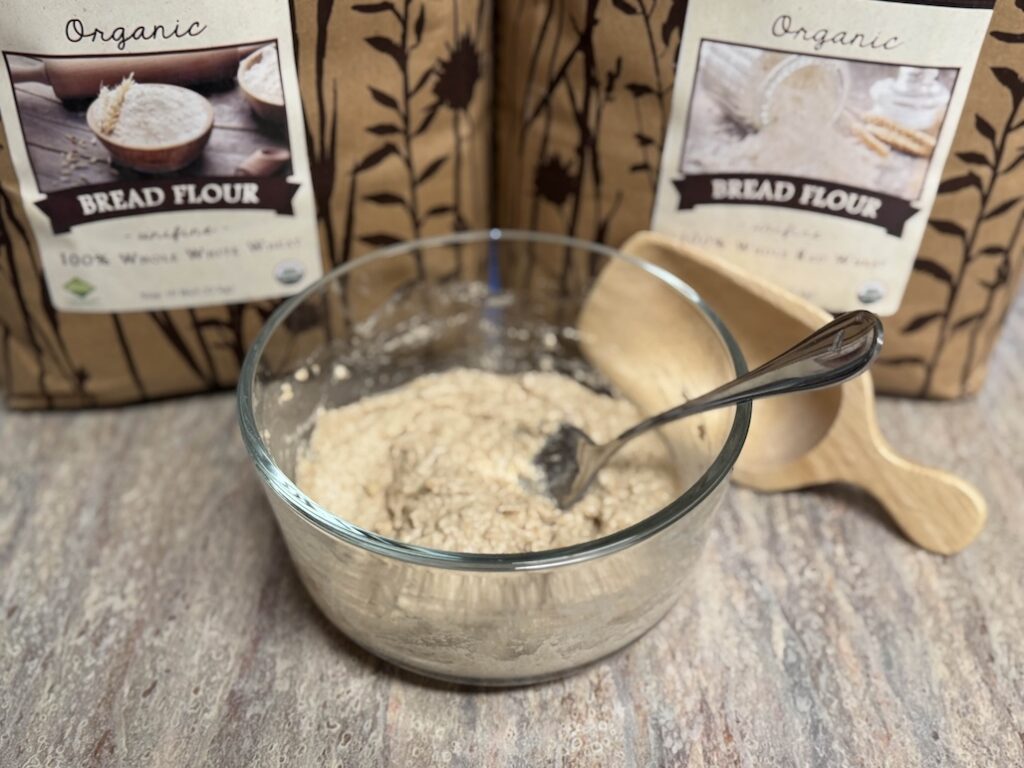 A glass bowl with sourdough levain in it and a silver spoon sticking out. Behind it is a wooden flour scoop and then two large bags of bread flour side by side