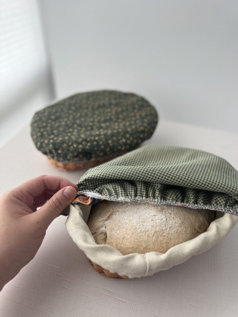 A hand is lifting off a green cotton bowl cover from a basket banneton that has whole wheat sourdough dough inside. In the background is another basket banneton that is covered with a sage green bread bowl cover