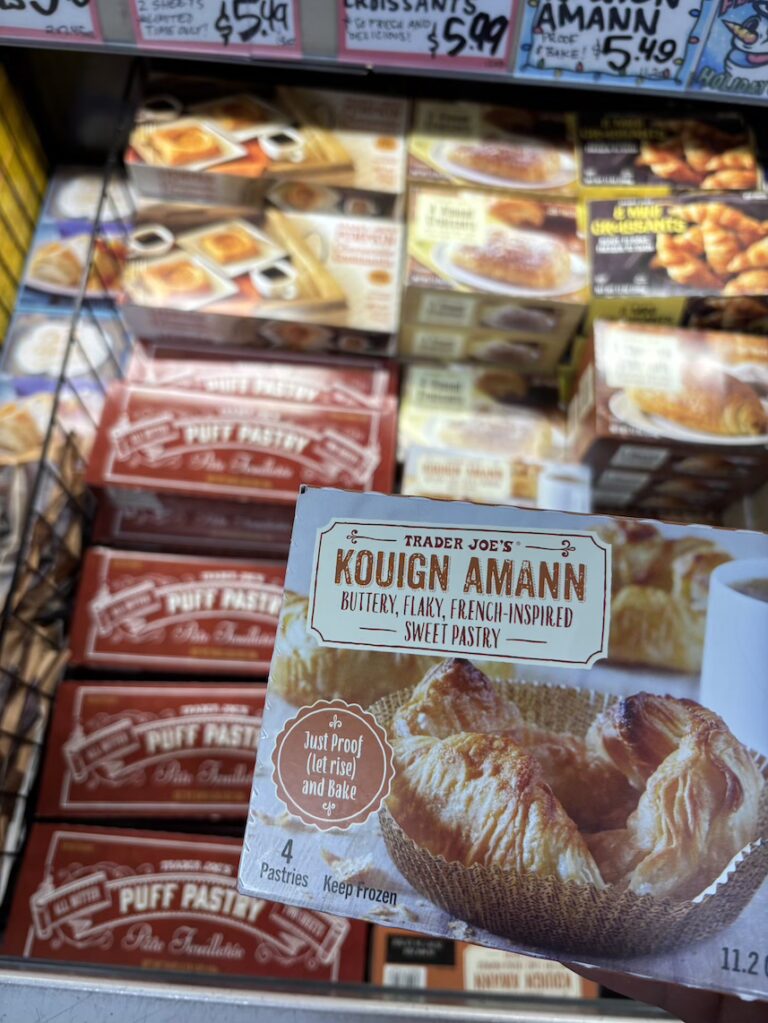 A hand holds a box of Trader Joe’s Kouign-Amann, described as a "Buttery, Flaky, French-Inspired Sweet Pastry". The box notes it contains 4 pastries and requires a "Proof (let rise) and Bake" process. There are various other frozen venoisserie options in the background.