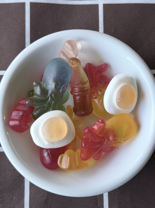 A top-down view of a variety of colorful French gummy candies in a white ceramic bowl. Visible shapes include a translucent teal octopus, classic gummy bears, and white-and-yellow fried egg gummies. The bowl sits on a dark brown tablecloth with a white grid pattern.