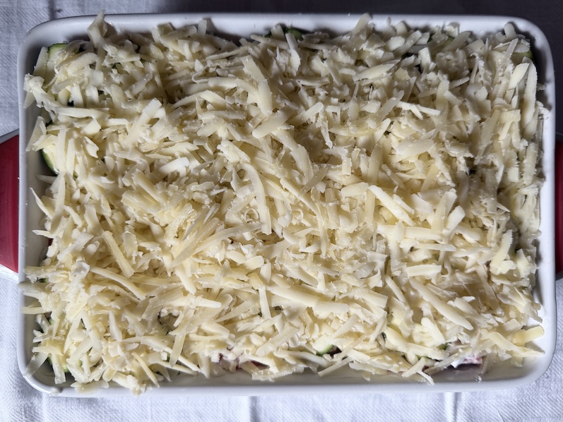 A thick, final layer of shredded Gruyère cheese completely covering the assembled salmon and zucchini gratin before baking.
