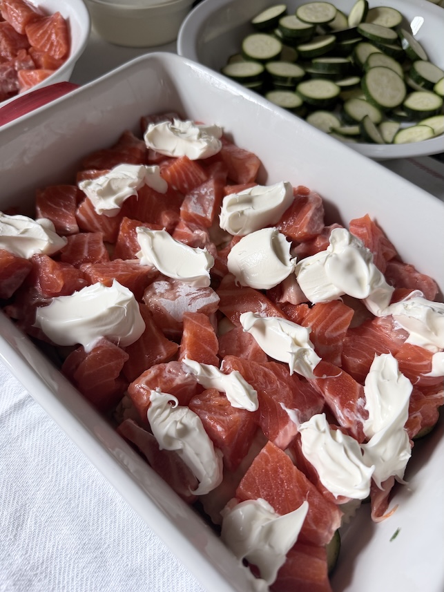 Dollops of thick crème fraîche placed evenly on top of cubed salmon pieces in a white casserole dish.