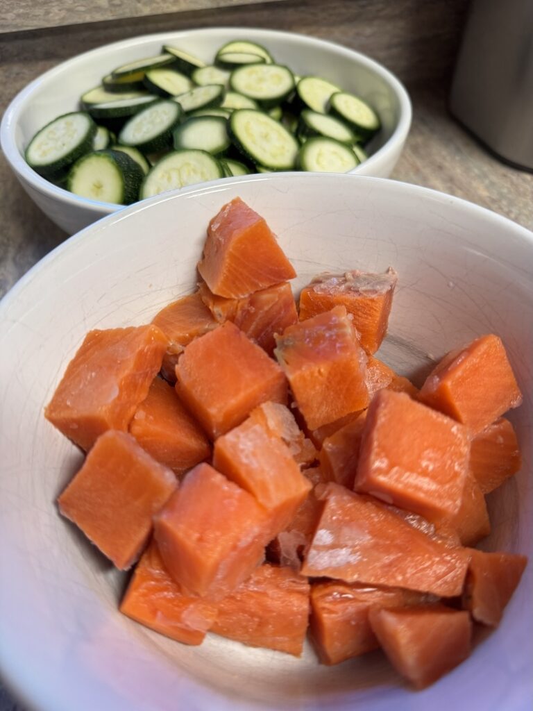 Fresh, bright orange cubed salmon pieces in a white bowl with a bowl of sliced green zucchini rounds blurred in the background.