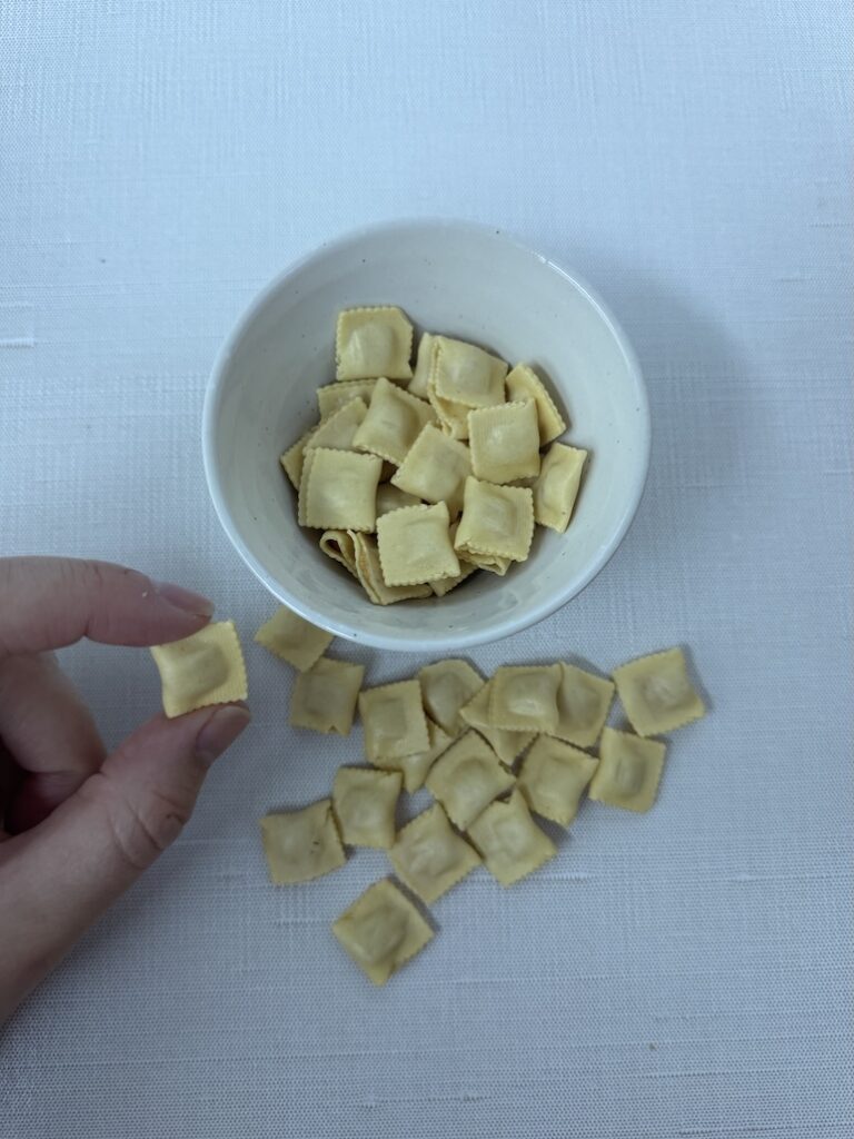 A hand holding a single piece of mini cheese ravioli over a small white bowl filled with uncooked micro ravioli pasta.