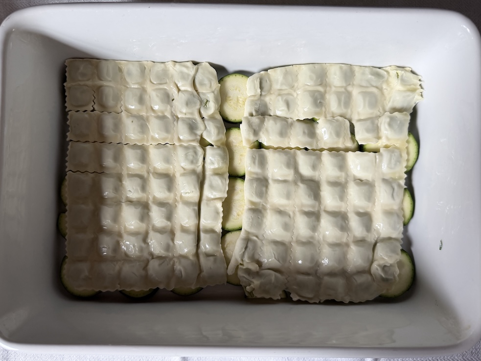 Top-down view of raviole pasta sheets layered flat over a base of fresh zucchini slices inside a ceramic baking pan.