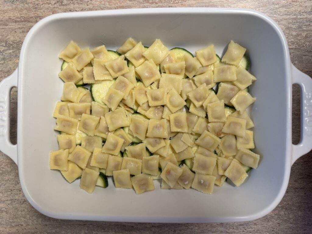 A layer of uncooked mini cheese ravioli scattered over sliced zucchini rounds in a white baking dish, showing the assembly process of a gratin.