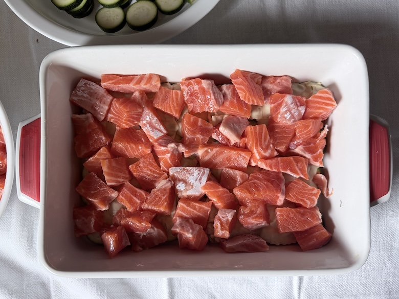 Adding a generous layer of raw cubed salmon pieces over the ravioli pasta during the assembly of a seafood gratin.