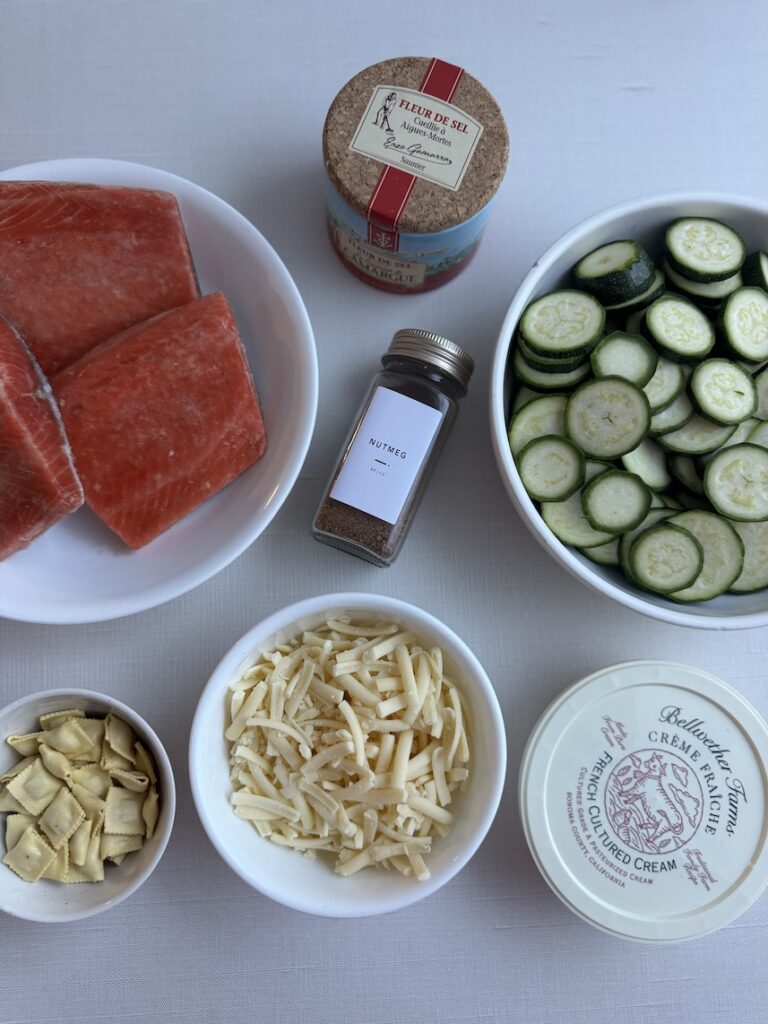Overhead flat lay of salmon zucchini gratin recipe ingredients, including raw salmon fillets, sliced zucchini, shredded cheese, crème fraîche, mini ravioli, nutmeg, and sea salt on a white surface.