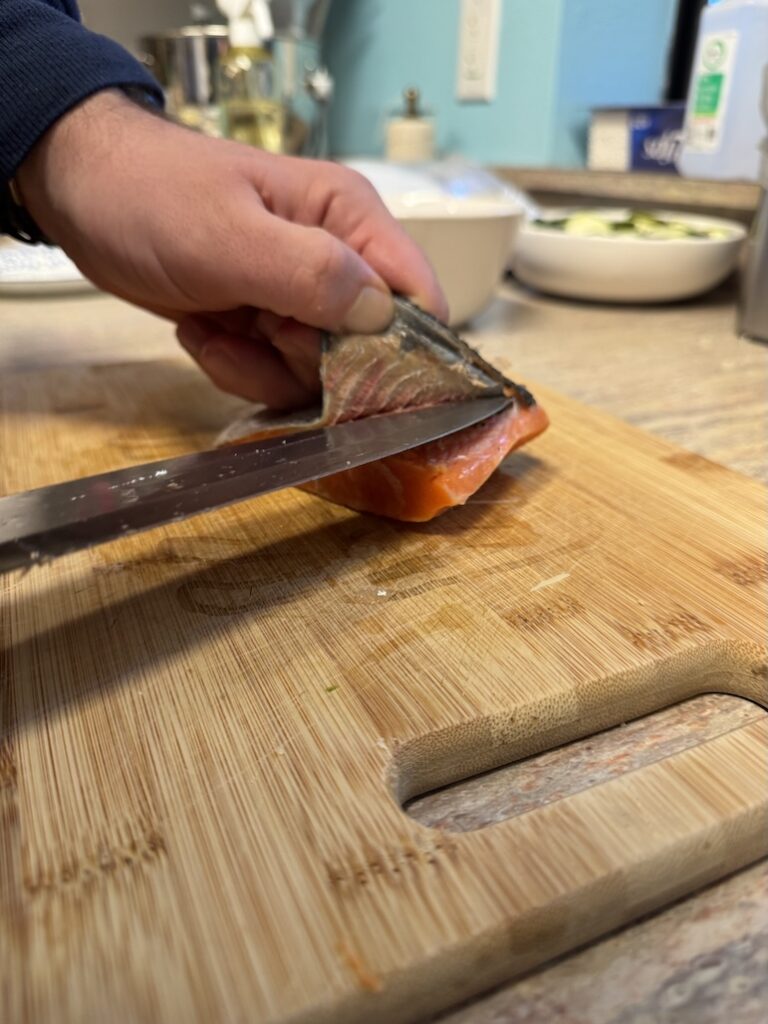Close-up action shot of a person using a sharp chef's knife to carefully skin a fresh salmon fillet on a wooden cutting board.