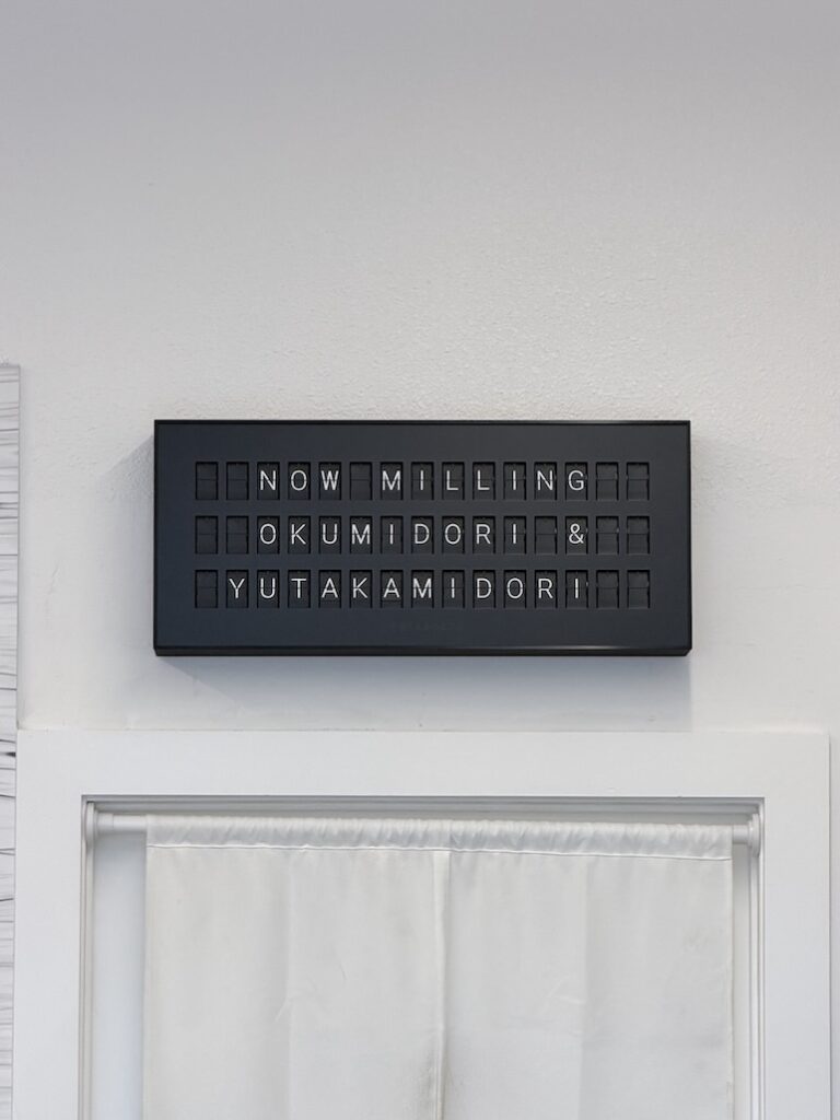 Minimalist black mechanical split-flap display reading "NOW MILLING OKUMIDORI & YUTAKAMIDORI" mounted above a white curtain.