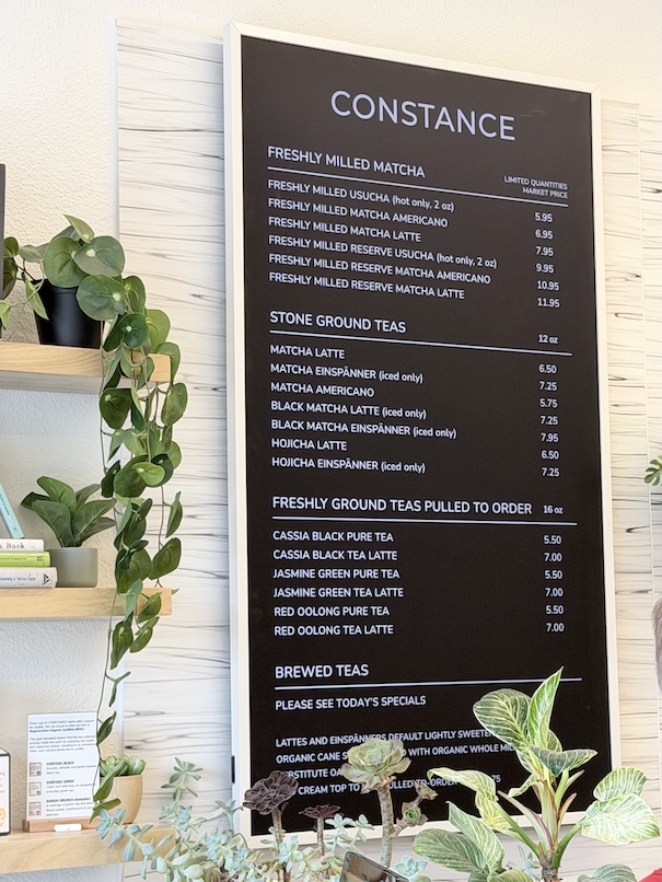 Black framed menu board at Constance Tea & Matcha listing prices for freshly milled matcha, stone ground teas, and brewed teas next to indoor plants.