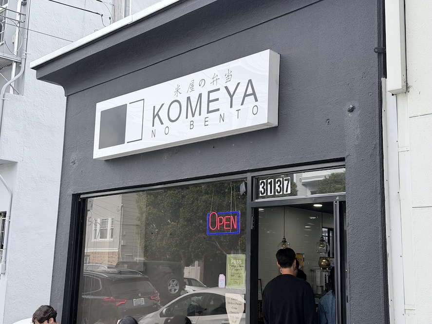 Exterior view of the Komeya No Bento Japanese restaurant storefront in San Francisco, featuring a dark gray facade and white logo sign above the entrance.