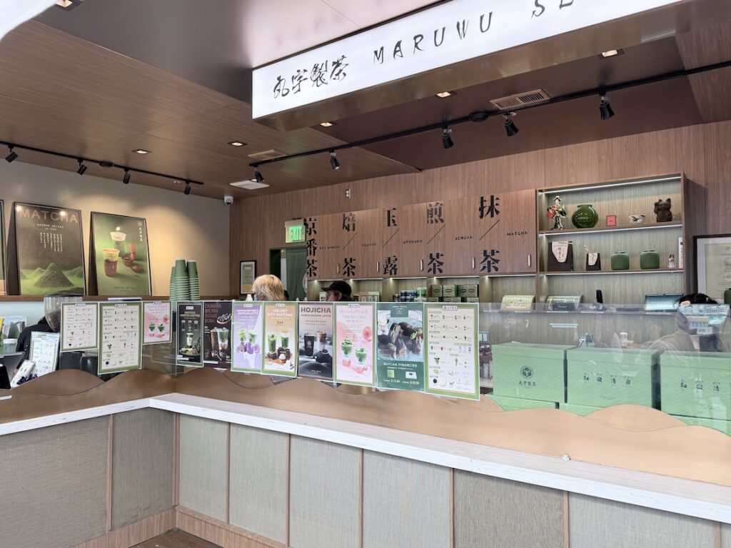 Wide angle interior shot of Maruwu Seicha tea shop showing the ordering counter, wood flooring, cheese tart promotional posters, and drink menu cards.