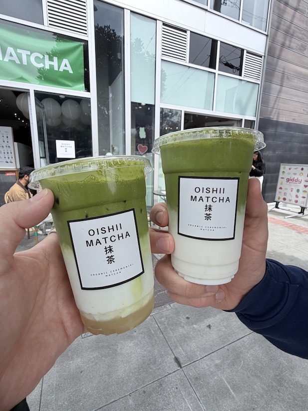 Two hands holding layered iced matcha lattes with milk in clear plastic Oishii Matcha cups outside the cafe.