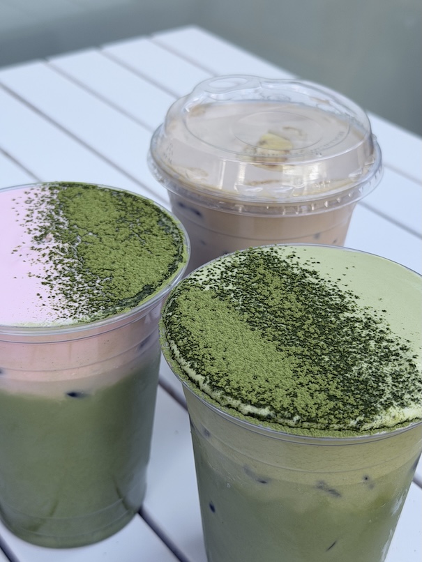 Three iced cafe drinks on a white table, featuring layered matcha lattes dusted with green matcha powder and a strawberry matcha top.